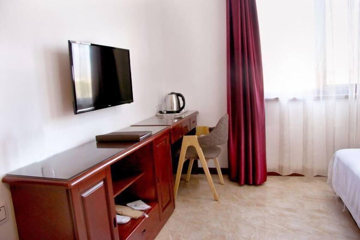 A hotel room with a flat-screen TV mounted on the wall, a wooden desk with a kettle, a chair, and a bed with white bedding.