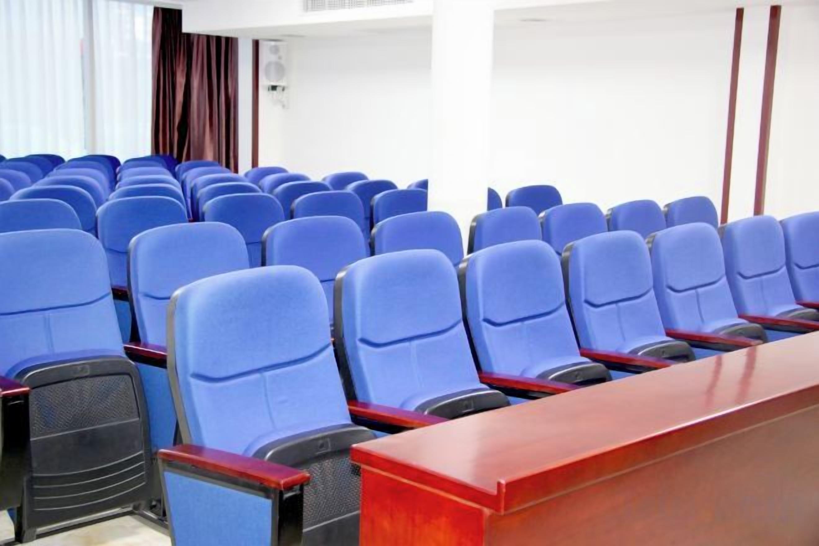Uma sala de conferências com fileiras de cadeiras azuis e uma mesa de madeira.