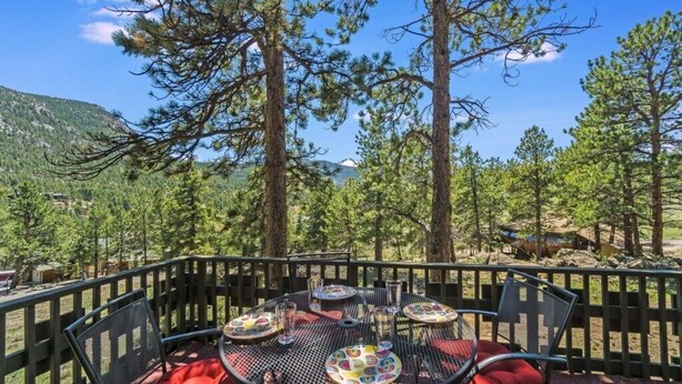 Eine Terrasse mit einem runden Tisch und Stühlen, umgeben von einem Holzzaun, mit Blick auf eine bewaldete Landschaft und Berge im Hintergrund.