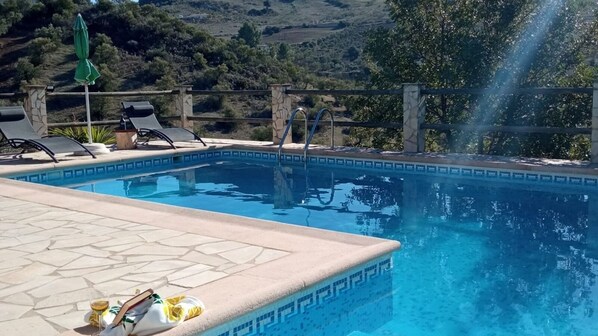 Piscina com borda de pedra, cadeiras de descanso e vista para as colinas.
