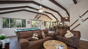 A spacious living room with a pool table, two brown leather sofas, a wooden coffee table, and a view of the backyard through large windows.