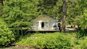 A riverside cabin with a deck overlooking a fast-flowing river, surrounded by dense greenery.