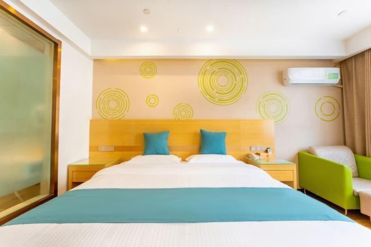 A hotel room with a large bed, two pillows, a bedside table, a green armchair, and a wall with circular patterns.