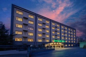 A multi-story hotel with a row of windows on each floor, a ground-level entrance, and a parking area in front.