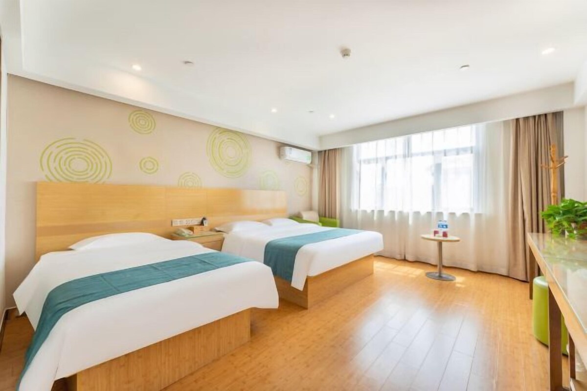 A hotel room with two beds, a bedside table, a small table with a lamp, and a window with curtains.