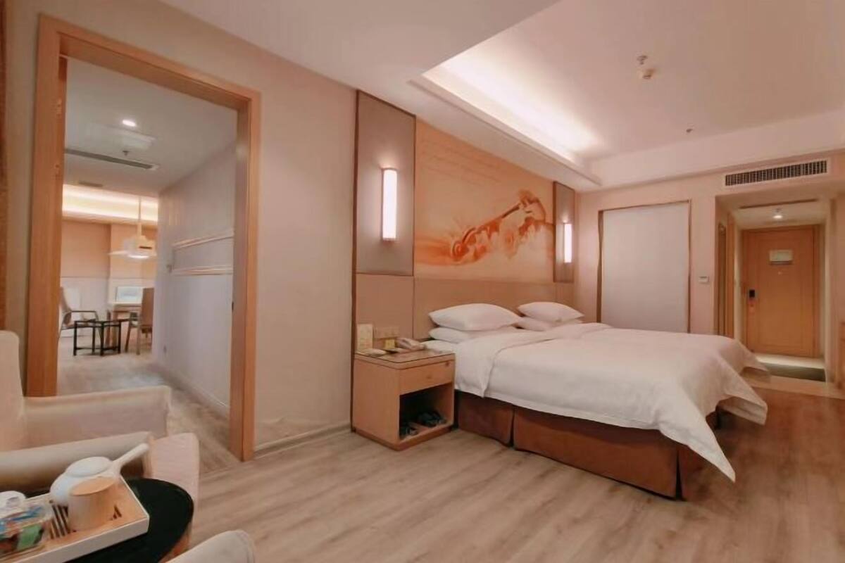 A modern hotel room with a large bed, a bedside table, a chair, and a desk with a tray of items.