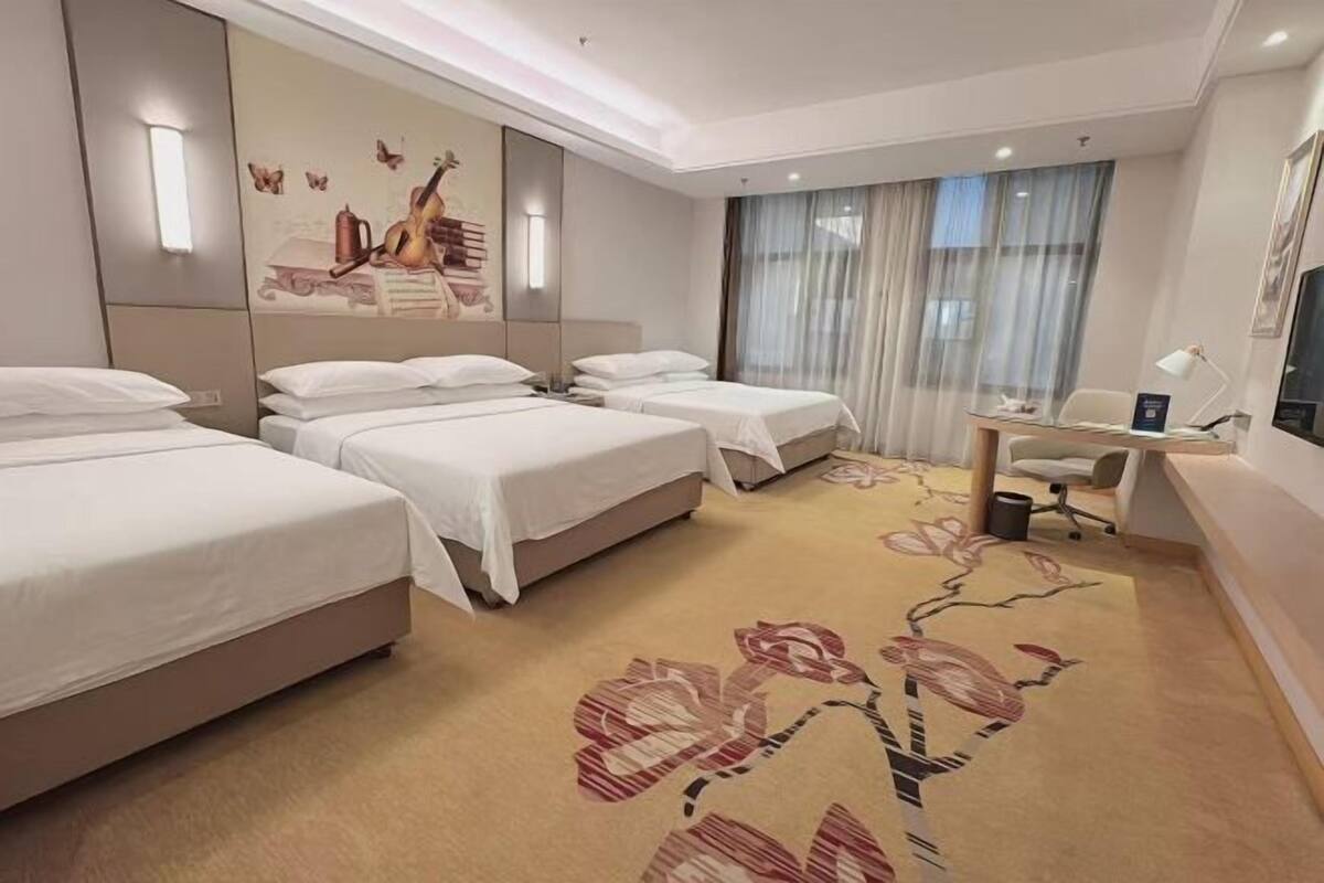 A hotel room with two beds, a desk, and a chair. There is a large window with curtains, a wall-mounted TV, and a decorative wall piece.