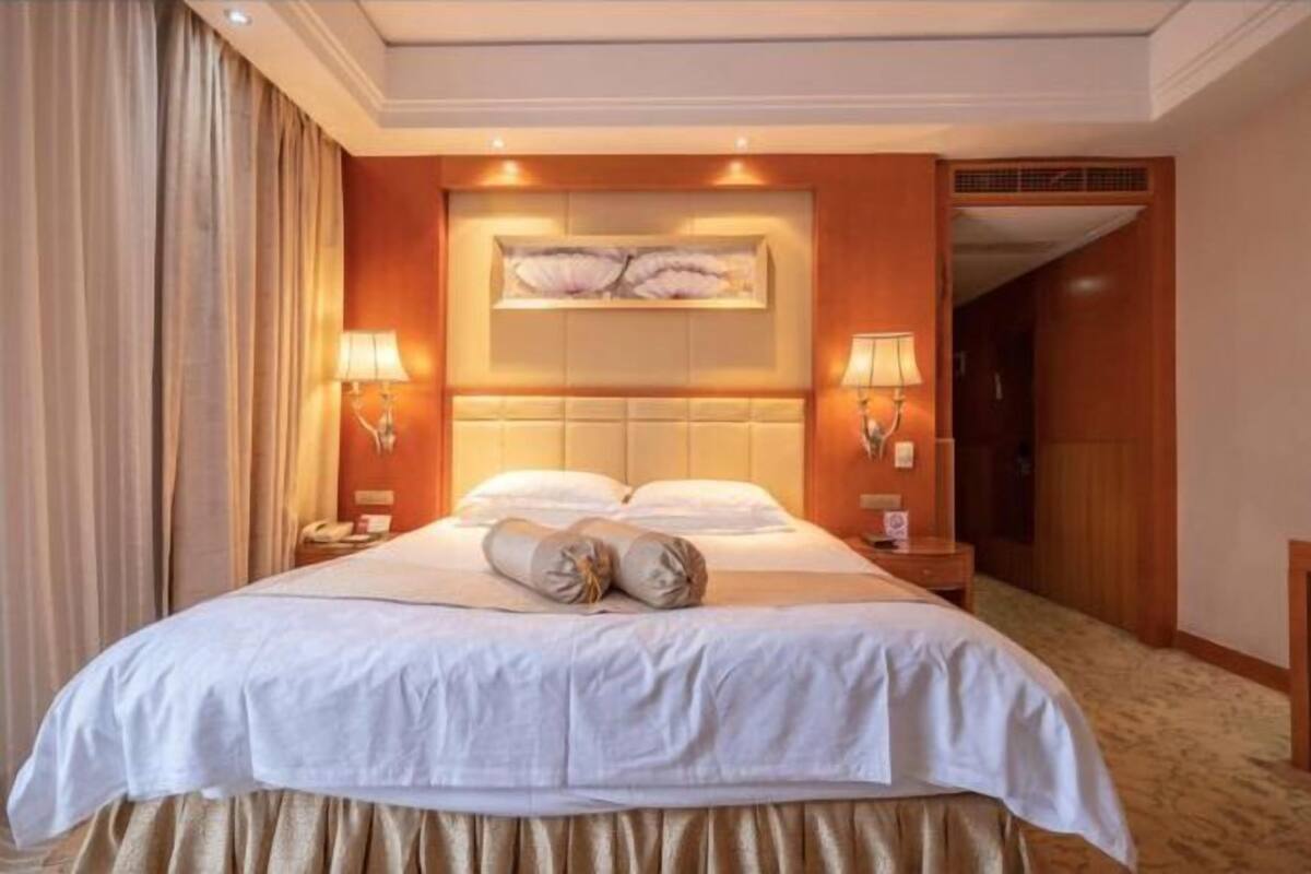 Hotellihuone, jossa on suuri sänky, kaksi yöpöytälamppua, kukka-aiheella koristeltu pääty ja matto lattialla.