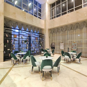 A spacious indoor area with a high ceiling, marble flooring, and a large bookshelf filled with books. There are multiple tables and chairs arranged for dining or socializing. - Vienna International Hotel (Shenzhen)