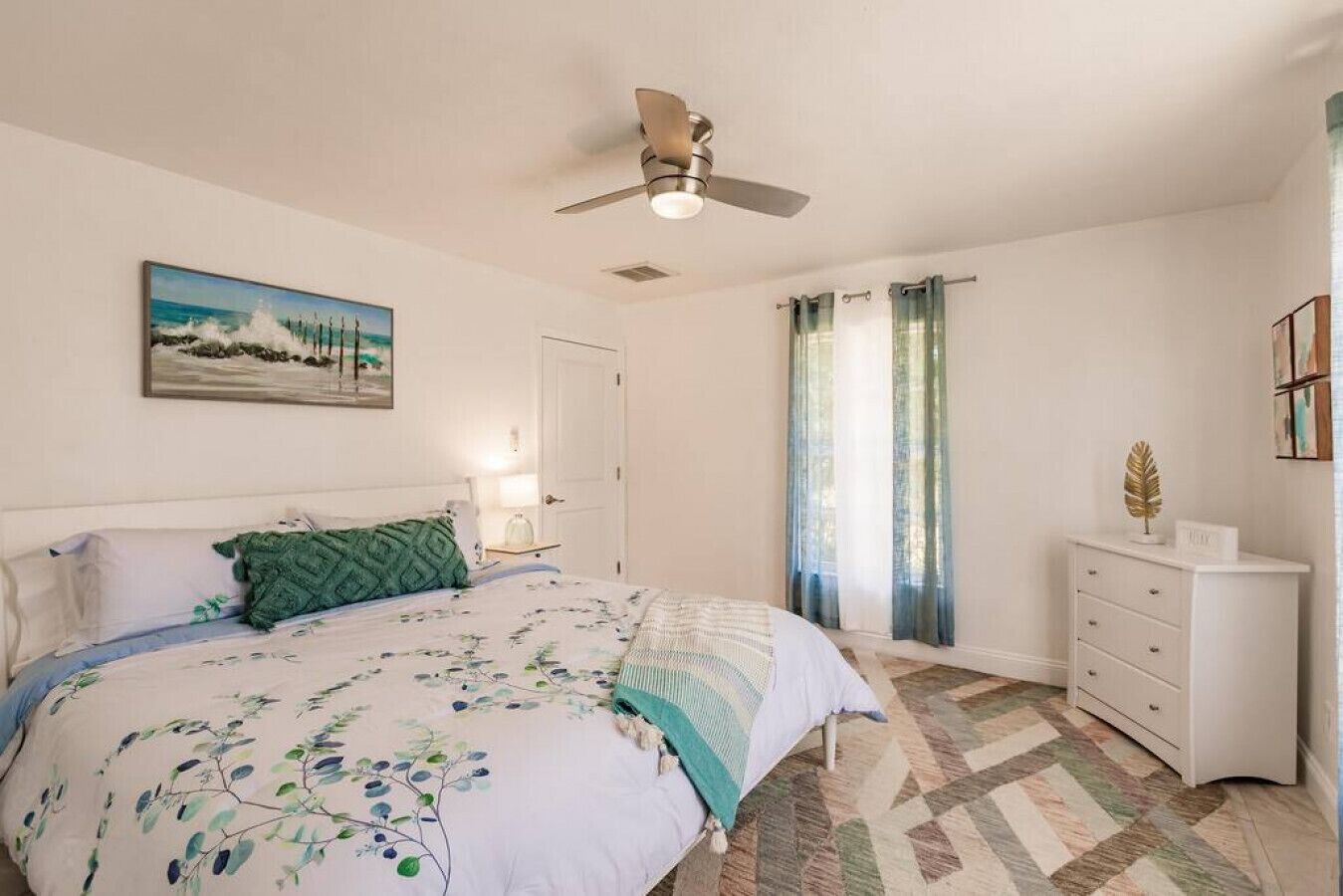 A bedroom with a bed, a ceiling fan, a nightstand, a dresser, and a painting on the wall.