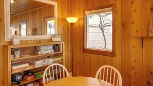 A wooden interior with a dining table, chairs, a shelf with board games, a lamp, and a window with a view of a tree. - Charming Lakefront Cabin w/ Deck & Fire Pit! (Newport)