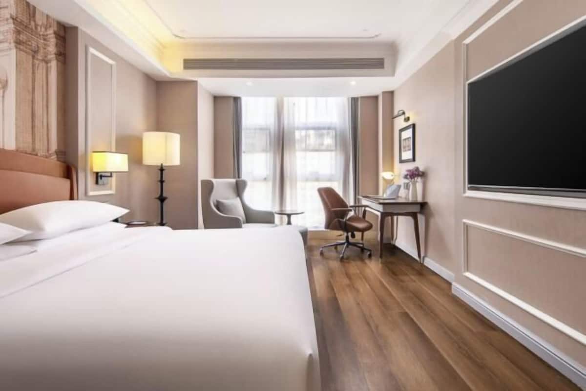 Une chambre d’hôtel avec un grand lit, un bureau, une chaise, une lampe et une grande fenêtre.