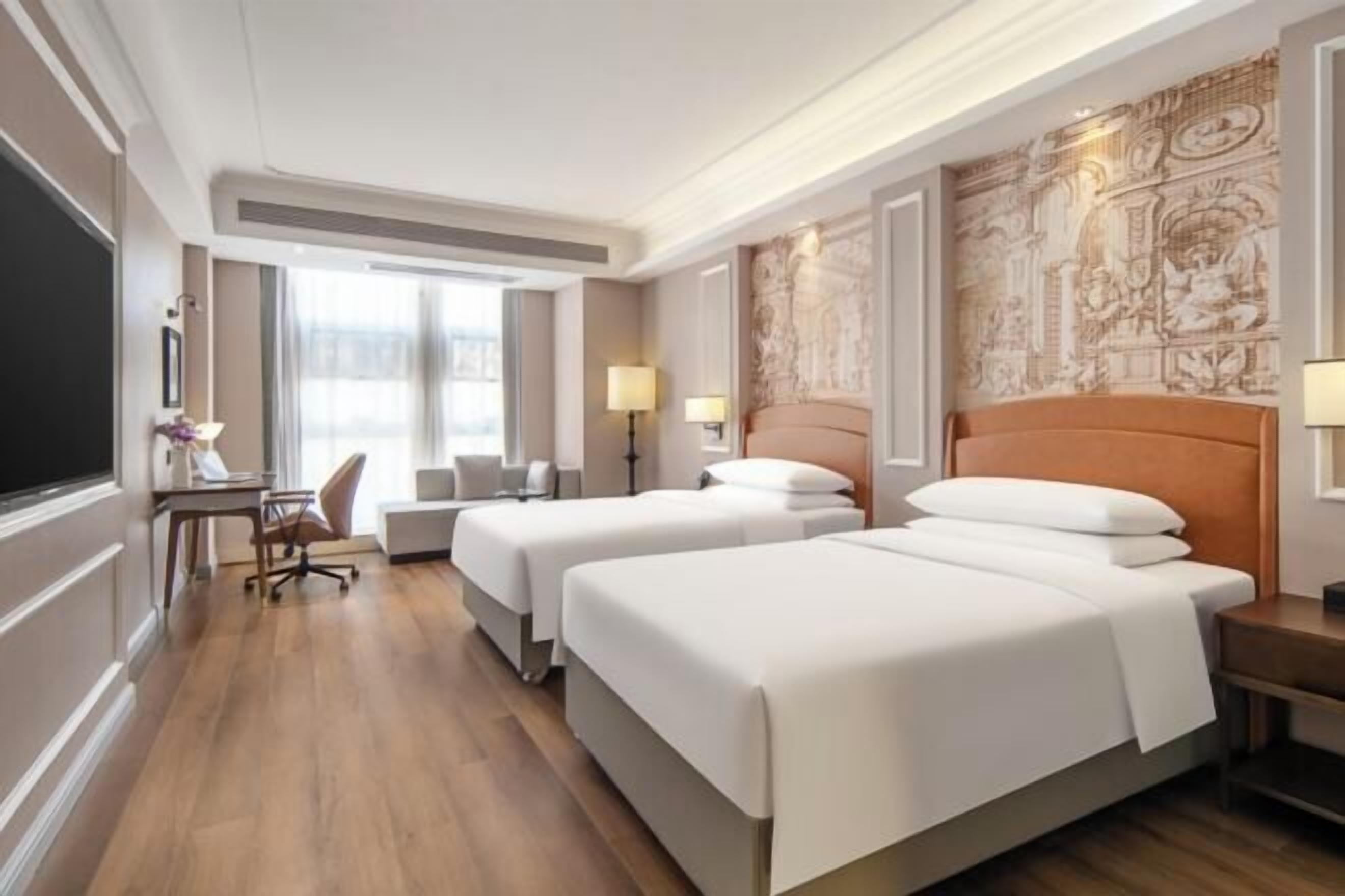 A hotel room with two beds, a desk, a chair, and a large window.