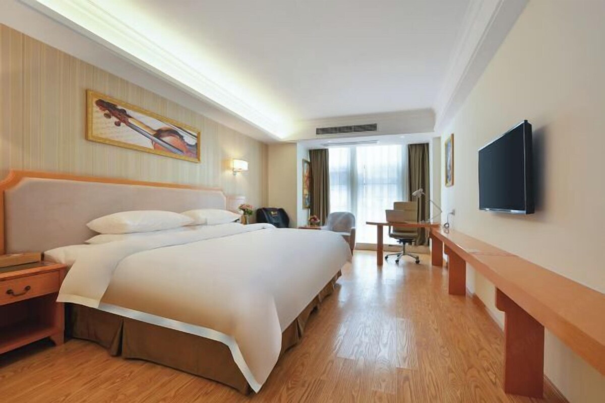 Ein Hotelzimmer mit einem großen Bett, einem Schreibtisch, einem Sessel, einem Fernseher und einem Bild an der Wand.