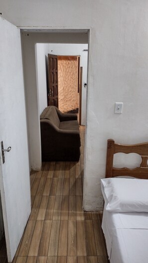 A room with a wooden floor, a bed with white bedding, and a sofa with a brown cover. - Quarto em Kitnet perto da primeira praia (Cairu)
