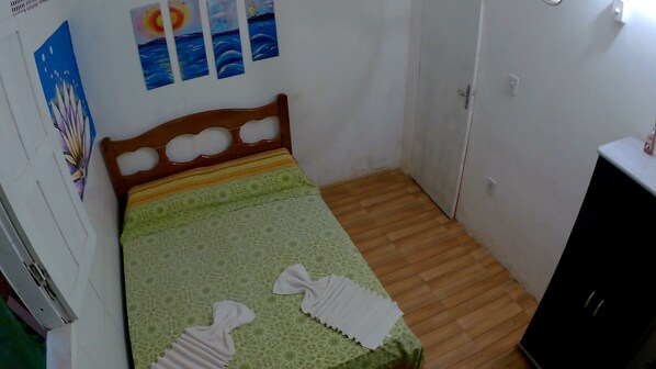 A small bedroom with a bed, a wooden headboard, a green patterned bedspread, and a wall with a painting of a sunset over the sea. - Quarto em Kitnet perto da primeira praia (Cairu)