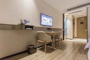 A hotel room with a bed, a desk, a chair, a TV mounted on the wall, a trash bin, and a door leading to another room.