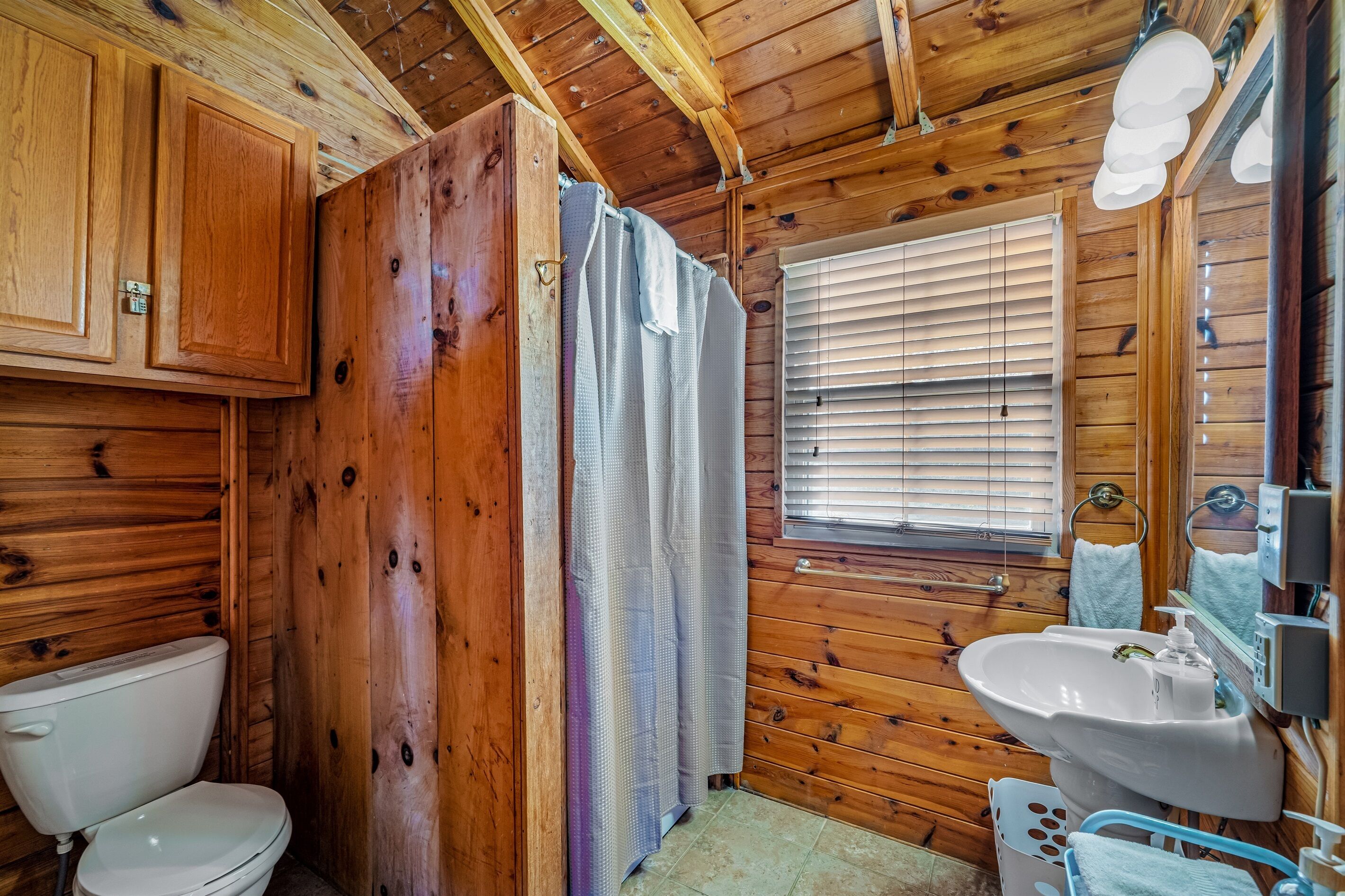 Een houten badkamer in een vakantiehuisje, met een toilet, wastafel en raam met jaloezieën.