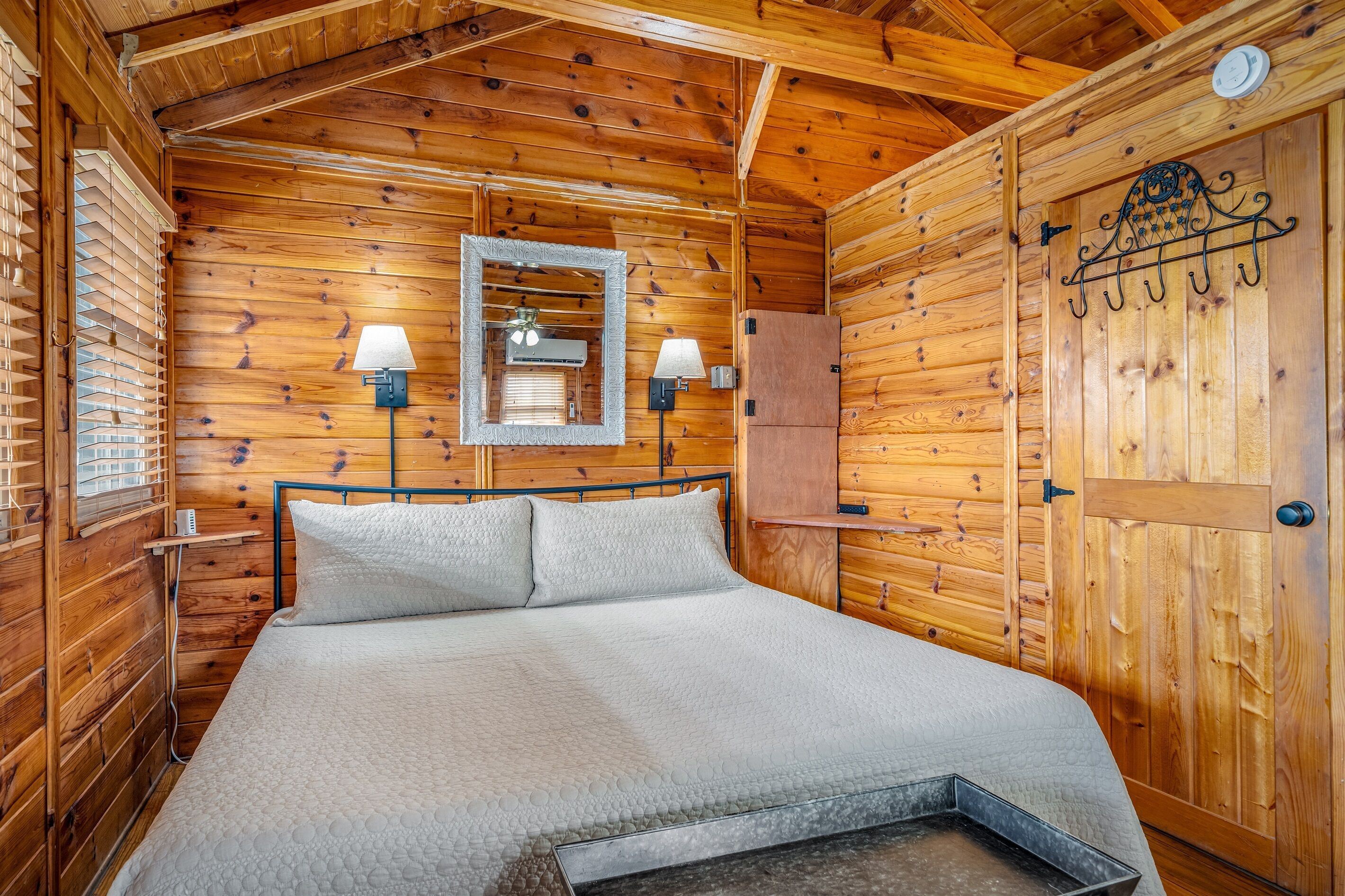 Ein Schlafzimmer einer Holzhütte mit einem Bett, zwei Wandleuchten, einem Spiegel und einer Tür mit Kleiderhaken.