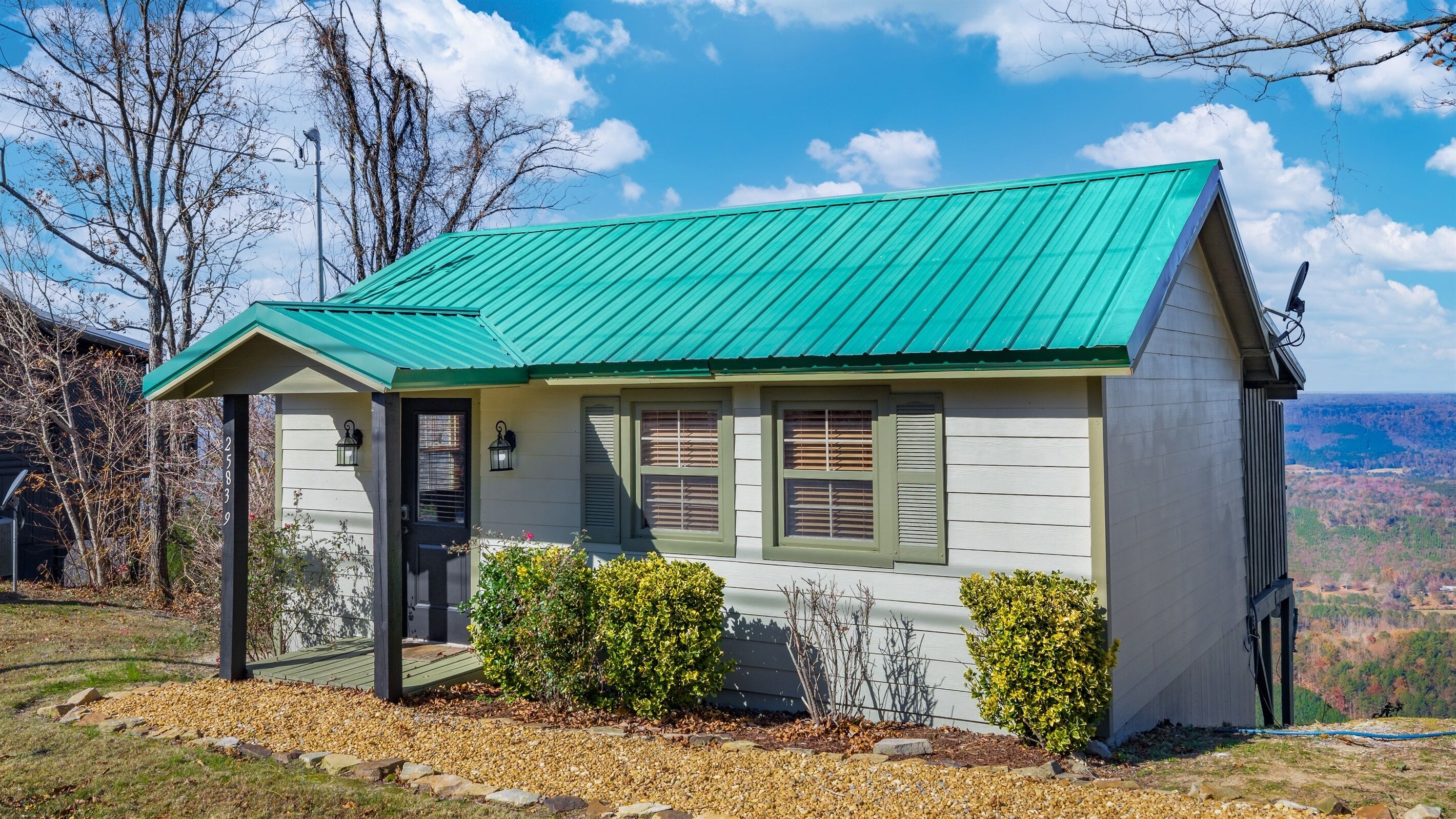 A small, single-story house with a green metal roof, a front porch, and a view of a mountainous landscape.