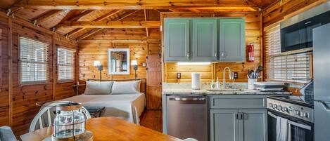 A log cabin interior with a wooden dining table, a bed, a kitchen with grey cabinets, and a large window.