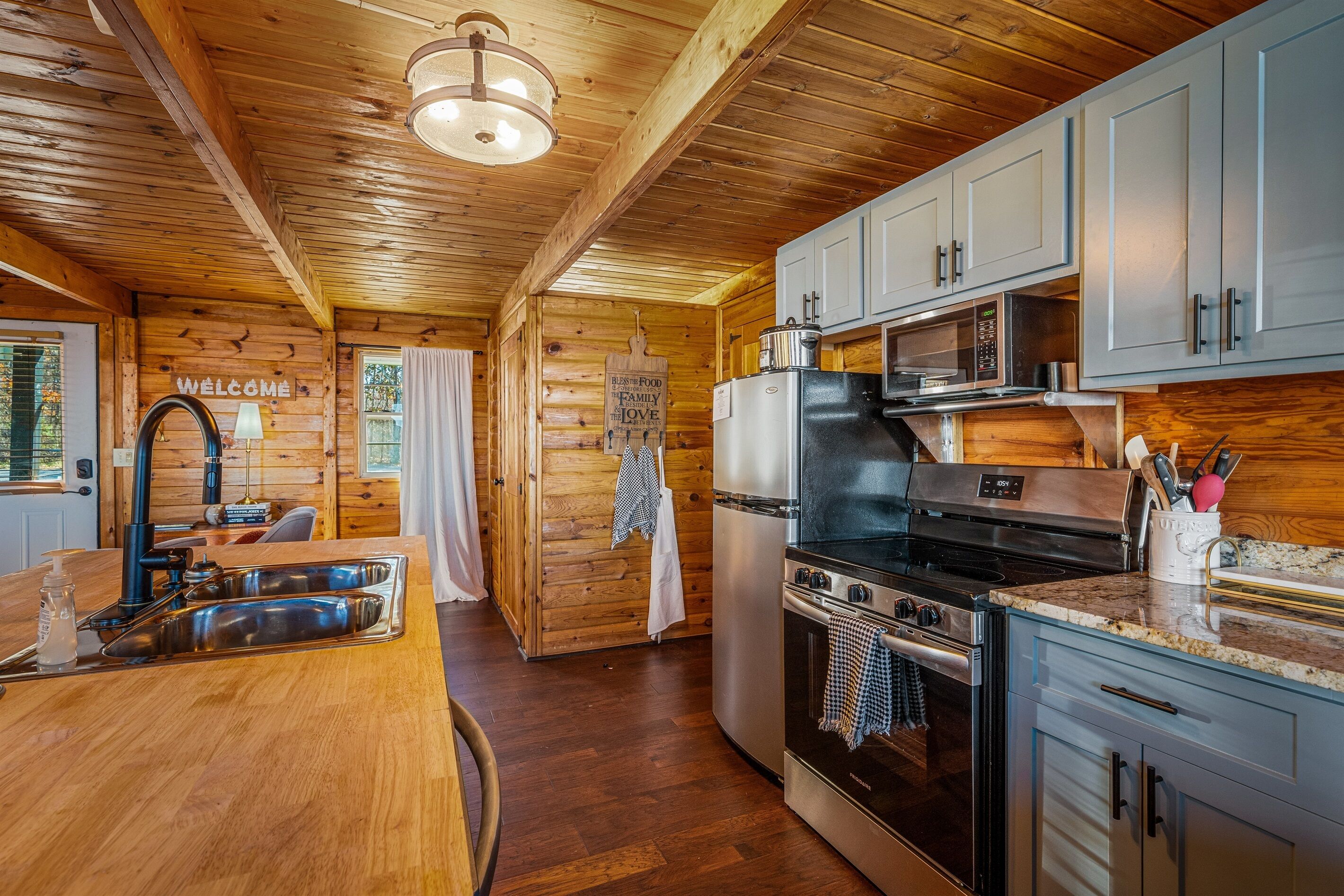 A cozy cabin-style kitchen with wooden walls, a stainless steel sink, a microwave, a stove, and cabinets.