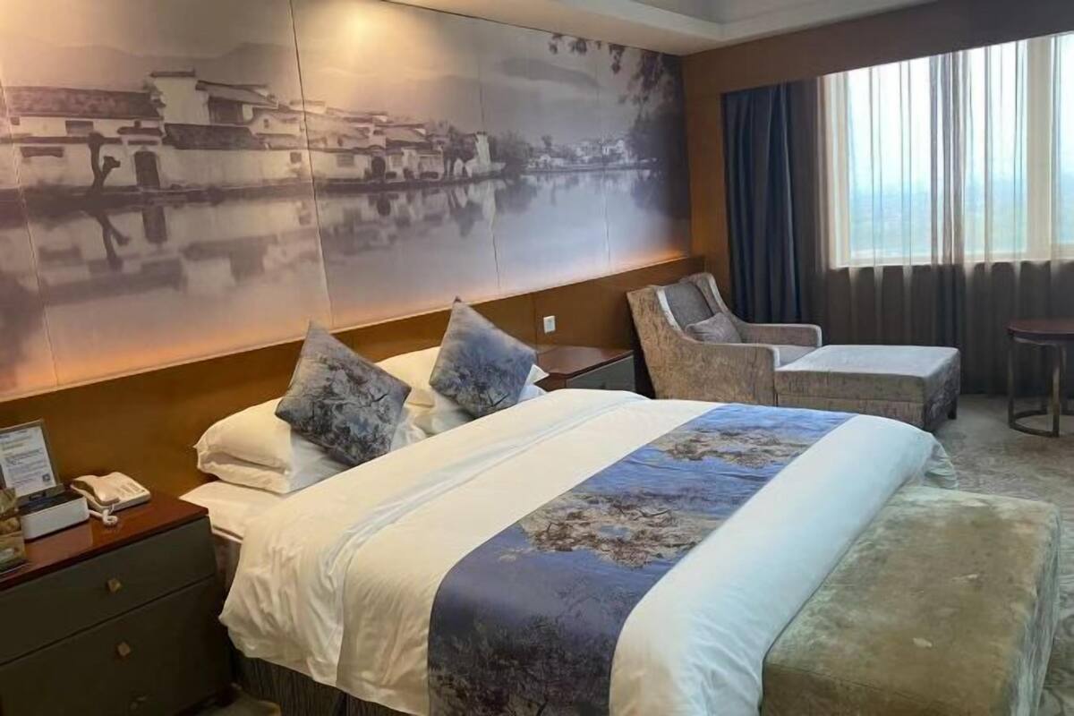 A hotel room with a bed, a chair, a small table, a nightstand, and a large wall mural.