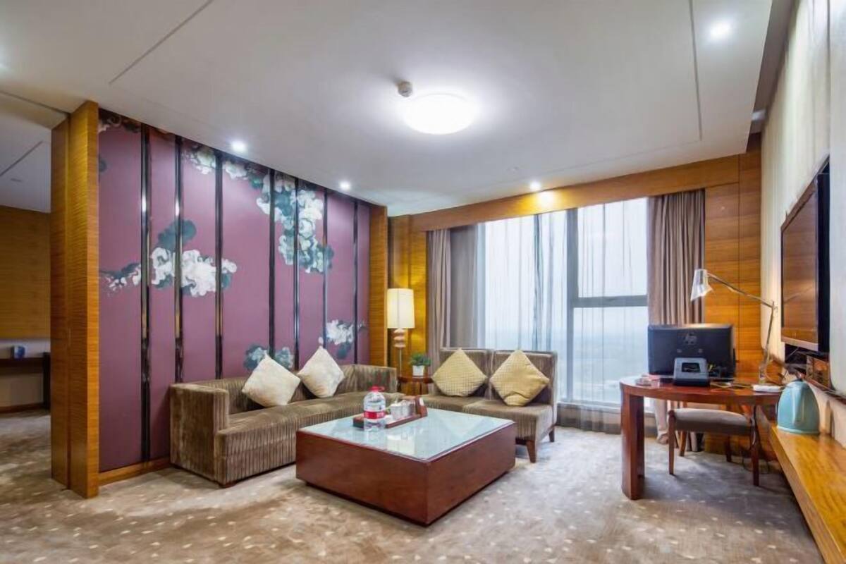 A hotel room with a sofa, a coffee table, a desk with a computer, a TV, and a large window with curtains.