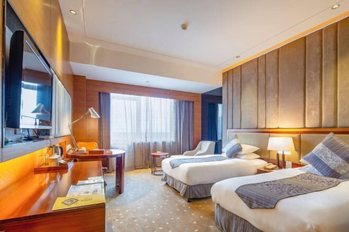 A hotel room with two beds, a desk, a chair, a TV, and a large window with curtains.