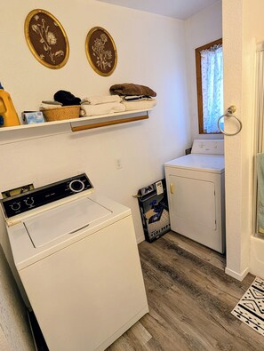 A laundry room with a front-loading washer and dryer, a shelf with folded clothes, and two framed wall decorations.
