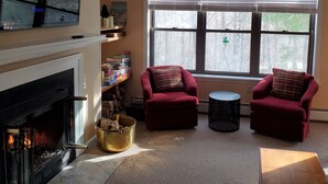 A cozy living room with a fireplace, two red armchairs, a coffee table, and a large window with a view of trees.