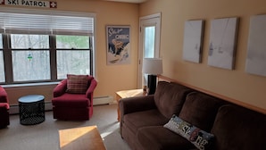 A cozy living room with a brown sofa, red armchair, and a wooden coffee table. A 'Ski Patrol' sign is visible on the wall, and a poster of a skier is also seen.