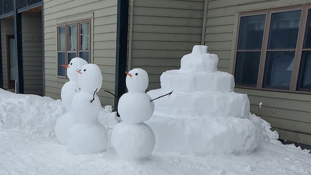 A snowman with a carrot nose and stick arms in front of a house.