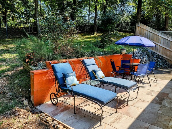 A patio with outdoor furniture, including lounge chairs and a table, surrounded by a wooden fence and a garden.