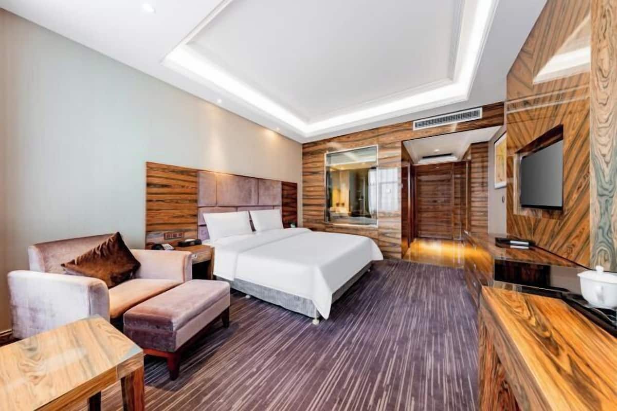 A modern hotel room with a large bed, a seating area with a chair and ottoman, a small table, a TV, and a wooden accent wall.