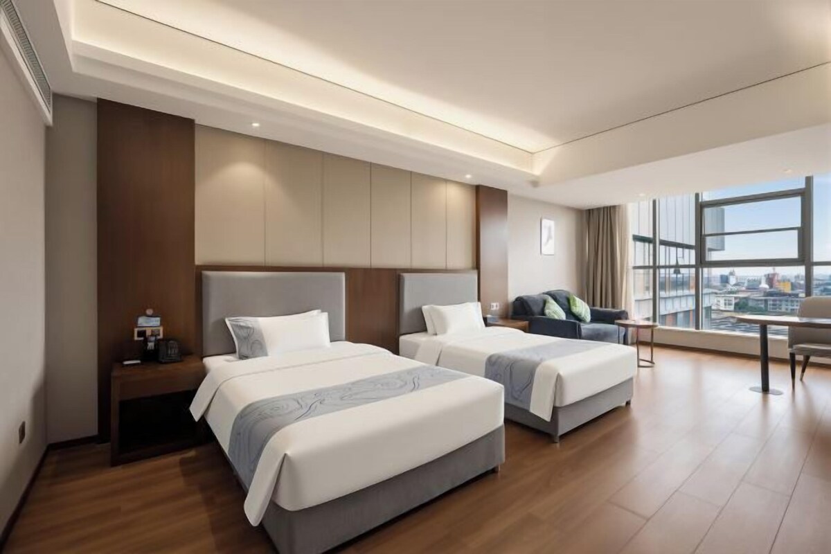 Een hotelkamer met twee bedden, een bureau en een groot raam met uitzicht op de stad.