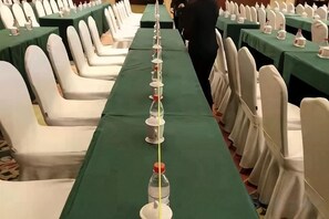 Een balzaal met rijen witte stoelen en tafels, elk met een groene tafelkleed en een waterfles.