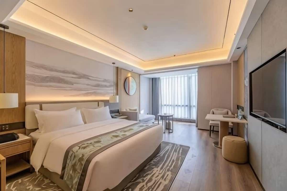 A modern hotel room with a large bed, bedside tables, a desk, a TV, and a large window.