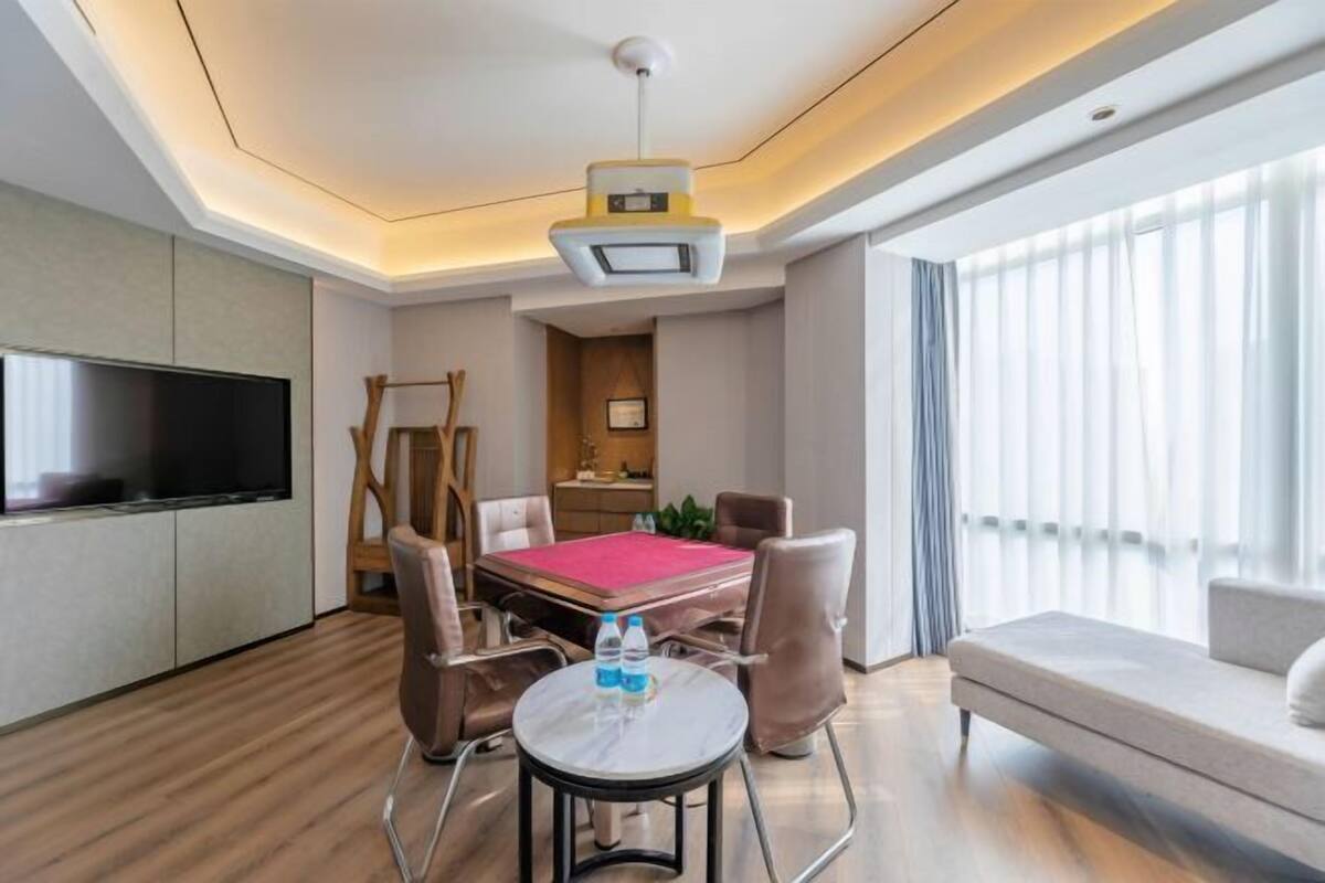 A hotel room with a flat-screen TV, a dining table with chairs, a sofa, and a large window with curtains.