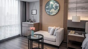 Una habitación de hotel moderna con sofá, una mesita con un florero, cama, mesita de noche, un cuadro en la pared y una ventana con cortinas traslúcidas.