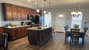 A spacious kitchen with wooden cabinets, a central island with stools, and a dining area with a chandelier. - Angler Sunrise..Luxury House on water (Hackberry)