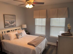 A bedroom with a bed, a ceiling fan, a nightstand, a lamp, and two windows with blinds.