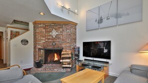 A living room with a brick fireplace, a flat-screen TV, and a cozy seating area. - Beautiful Fall Line Condo with Mountain Views on Shuttle Route B4A/B (Killington)