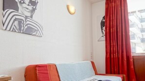 A room with a wooden sofa bed, a wall-mounted light, and a window with red curtains. - Lovely apartment for 2 people with WIFI (Les Boisses)