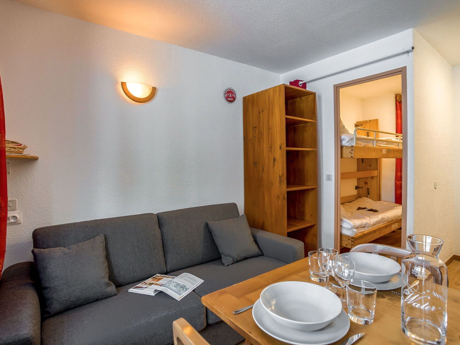 Une pièce de taille modeste avec un canapé gris, une table en bois sur laquelle se trouvent des assiettes et des verres, une armoire en bois et, au fond, un lit superposé.