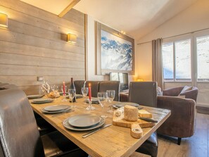 Uma sala de jantar com uma mesa de madeira posta para quatro pessoas, incluindo pratos, copos e talheres. Uma tábua de queijos com um queijo inteiro e uma garrafa de vinho também são visíveis. O ambiente possui painéis de madeira e uma grande janela com vista para montanhas.