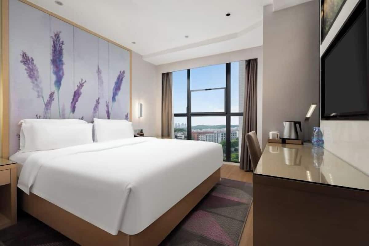 A hotel room with a large bed, a desk, a view of the city, and a wall with floral artwork.