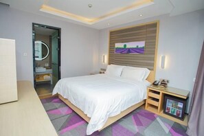 A modern hotel room with a large bed, a bedside table with a phone and a small fridge, a mirror, and a view of a field through the window.