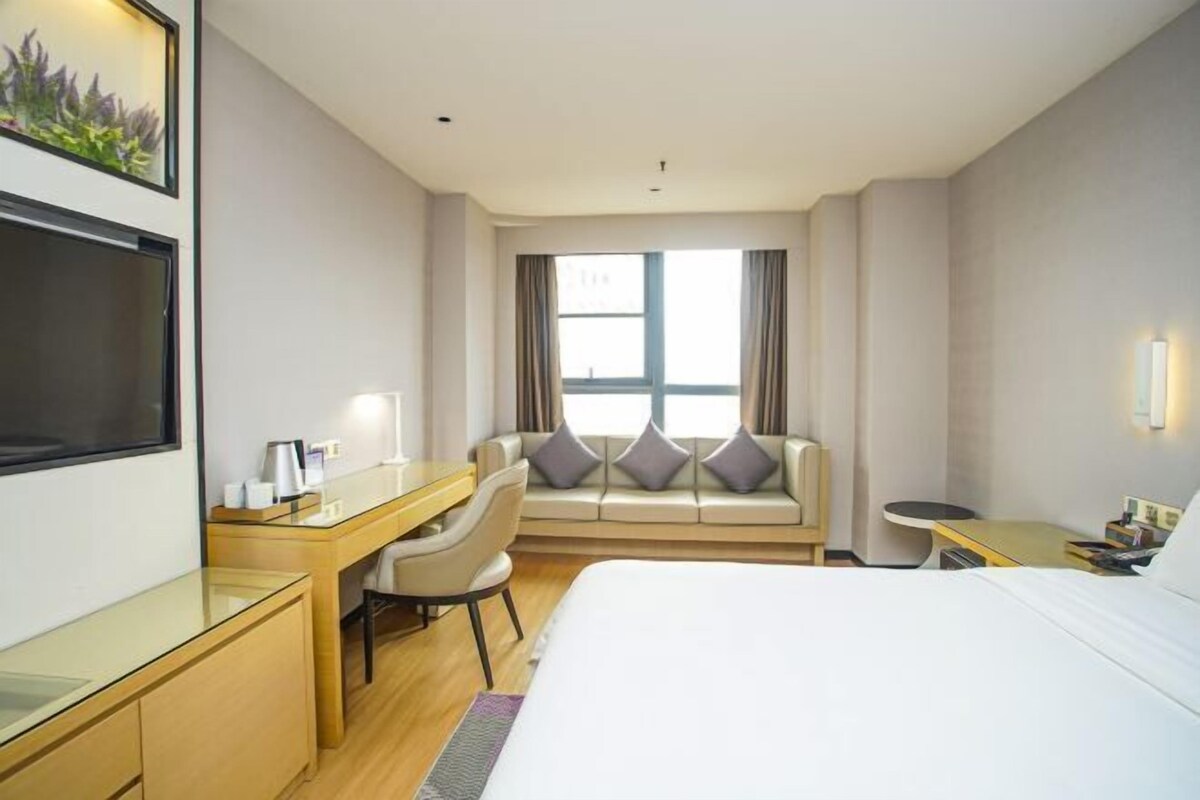 A hotel room with a bed, a desk, a chair, a sofa, a window with curtains, and a wall-mounted TV.
