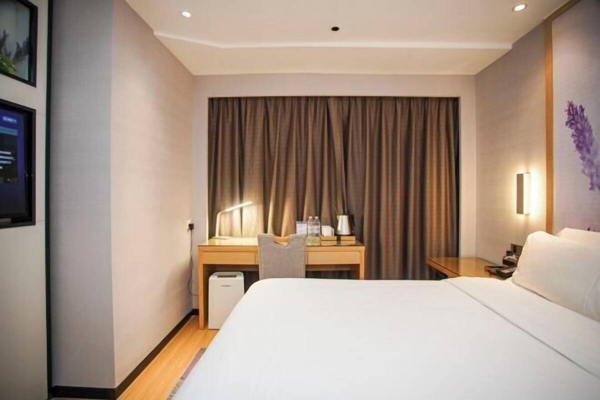 A hotel room with a large bed, a desk with a chair, a desk lamp, a nightstand, and a wall-mounted TV.